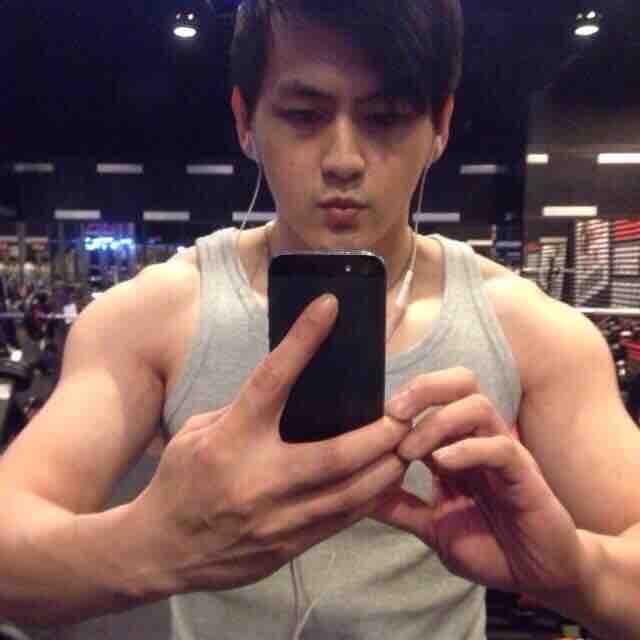 這張照片是一個年輕男子在健身房自拍。他穿著灰色背心,展示了他健壯的肌肉,並用手機自拍。他看起來很自信,也很享受健身的過程。