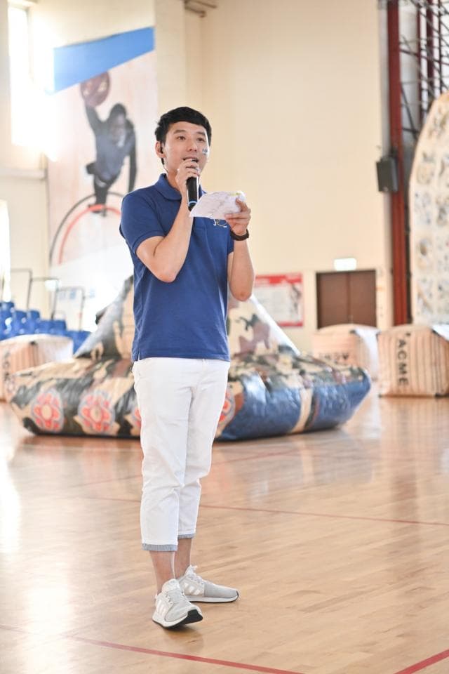 這張「男人正在體育館裡演講」的照片是,他穿著藍色的襯衫和白色的褲子,拿著麥克風,站在一個充氣的遊戲場地前面。