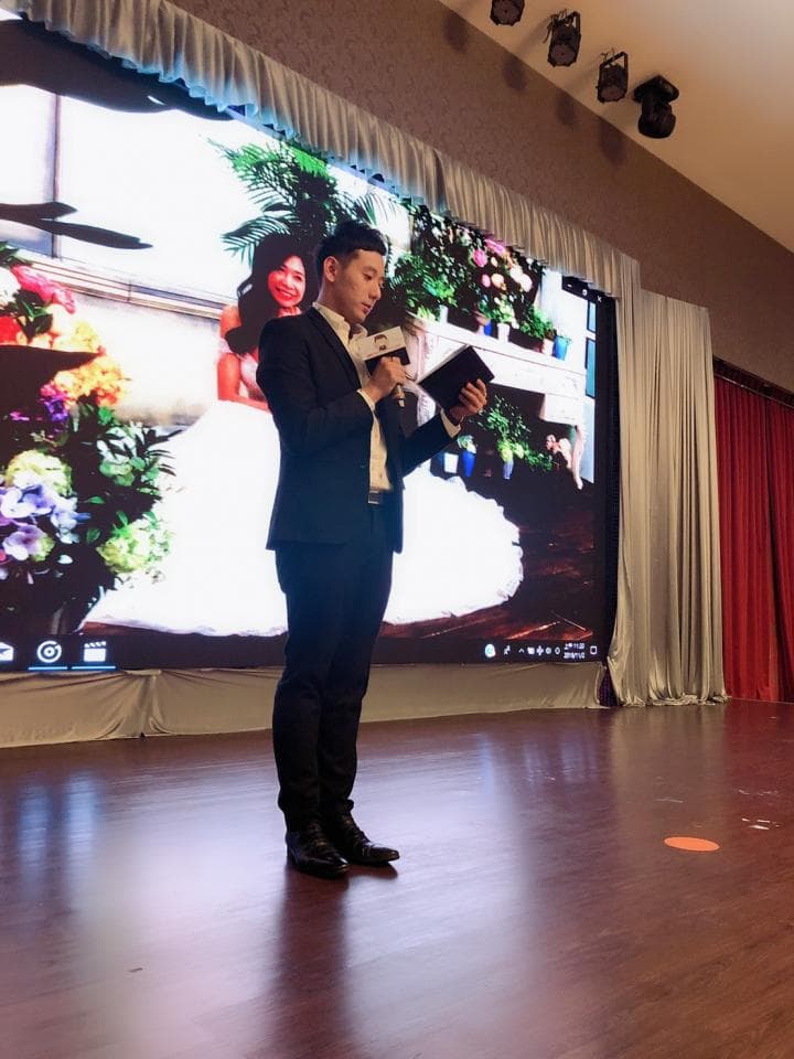 這張「婚禮」的照片是一位穿著西裝的男人正在臺上講話,他手中拿著麥克風和一個文件夾。