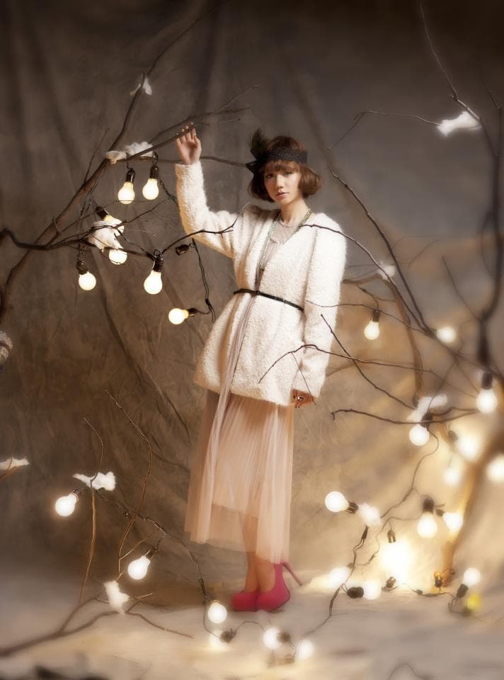 這張「一個穿著白色外套和粉色裙子的女孩站在樹枝上,樹枝上掛著許多燈泡」的照片是。