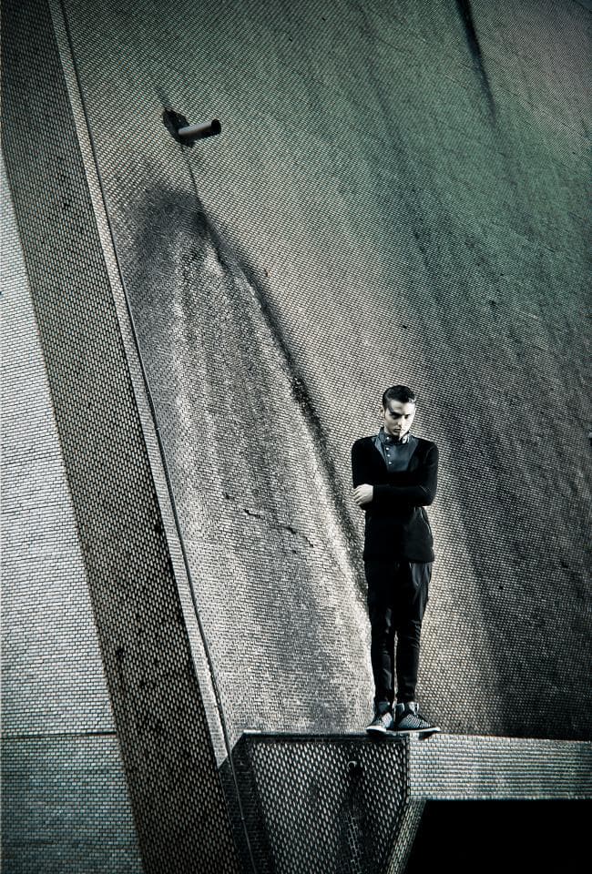 這張照片是一個男人站在水泥牆上,他穿著黑色衣服,雙手交叉抱胸,他的表情看起來很憂鬱。牆上有一條鐵管,背景是水泥牆。