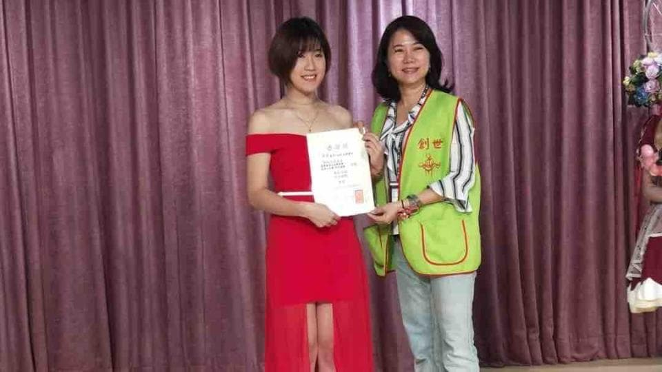 這張照片是一名穿著紅色禮服的年輕女性正在接受一位穿著綠色背心的女性頒發證書。