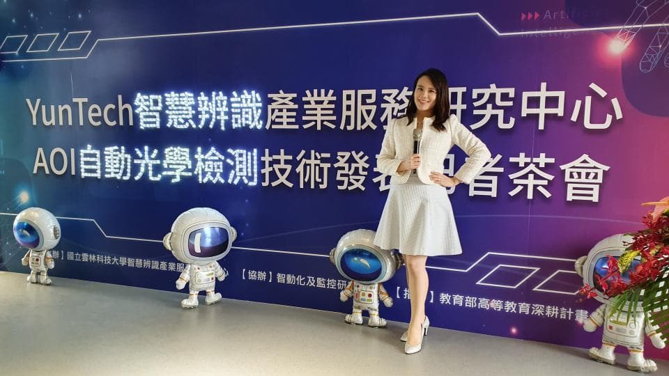 這張「科技公司活動」的照片是,一名女性站在一個藍色背景牆前,牆上寫著「YunTech 智識 辨識 產業服務研發中心 AOi 自動光學檢測技術研發 臺灣茶會」,背景牆上還有幾個機器人玩偶。