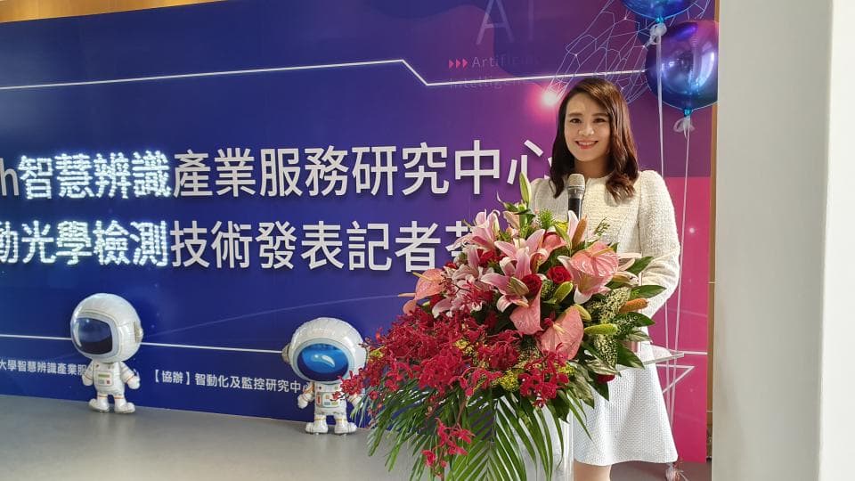 這張照片是一個女性站在講臺上,她手持麥克風,身後有一束鮮豔的花束,她的背後是一塊藍色的背景板,上面寫著「智慧聯網產業服務研究中心」。