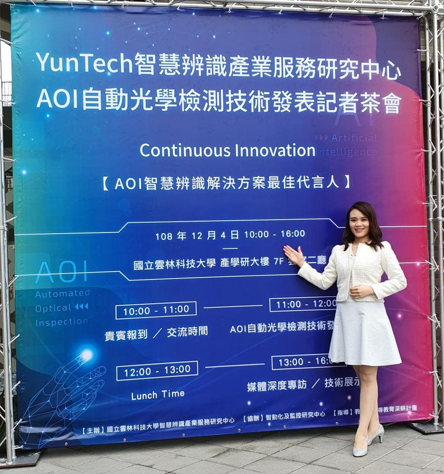 這張「雲科技智慧辨識產業服務研究中心AOI自動光學檢測技術研討會」的照片是,一個穿著白色洋裝的女士,站在一塊寫著「雲科技智慧辨識產業服務研究中心AOI自動光學檢測技術研討會」的藍色布條前面。