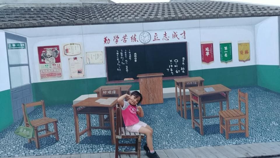 這張「教室」的照片是一個畫在牆上的教室場景,裡面有書桌、椅子和一個黑板。一個小女孩坐在一張書桌旁,看起來很開心。