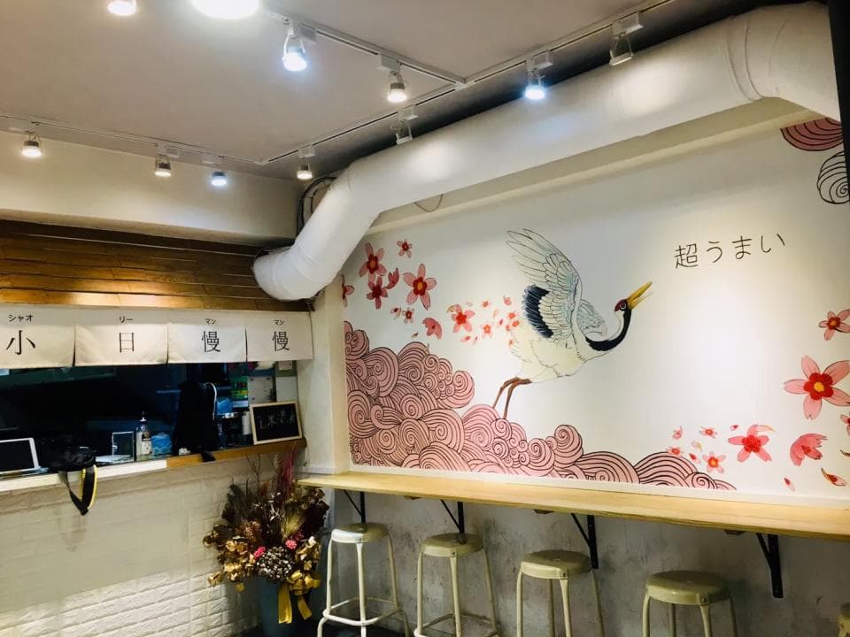 這張「餐廳」的照片是牆壁上有一隻畫著的鶴,還有粉紅色的花朵,以及日文漢字。牆壁的顏色是白色,牆上掛著燈,旁邊有木板做成的桌子和椅子。