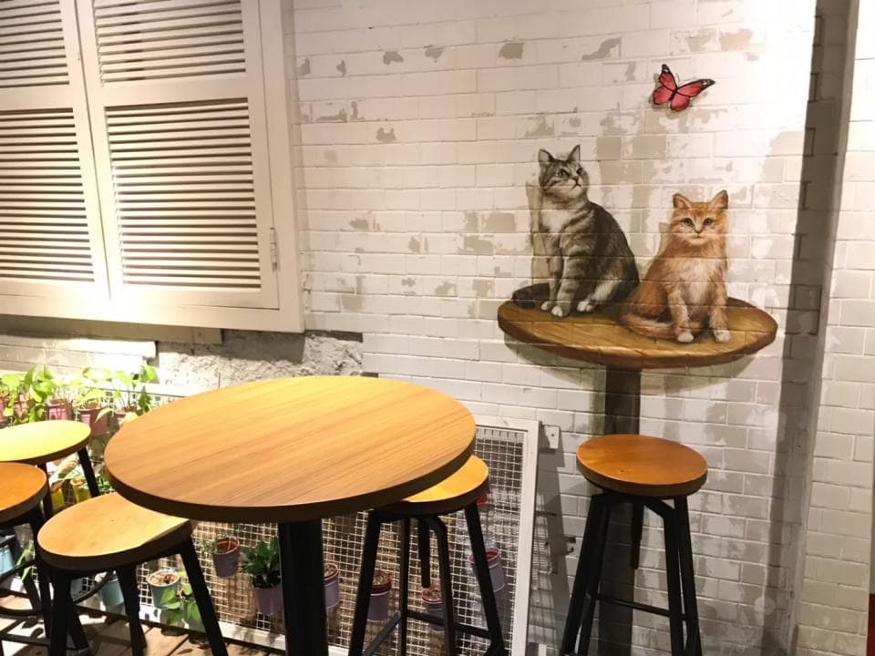 這張「貓咪壁畫」的照片是一間咖啡廳,牆上畫著兩隻可愛的貓咪,一隻灰色的,一隻橘色的,還有蝴蝶。咖啡廳裡擺放了幾張木桌和高腳椅。