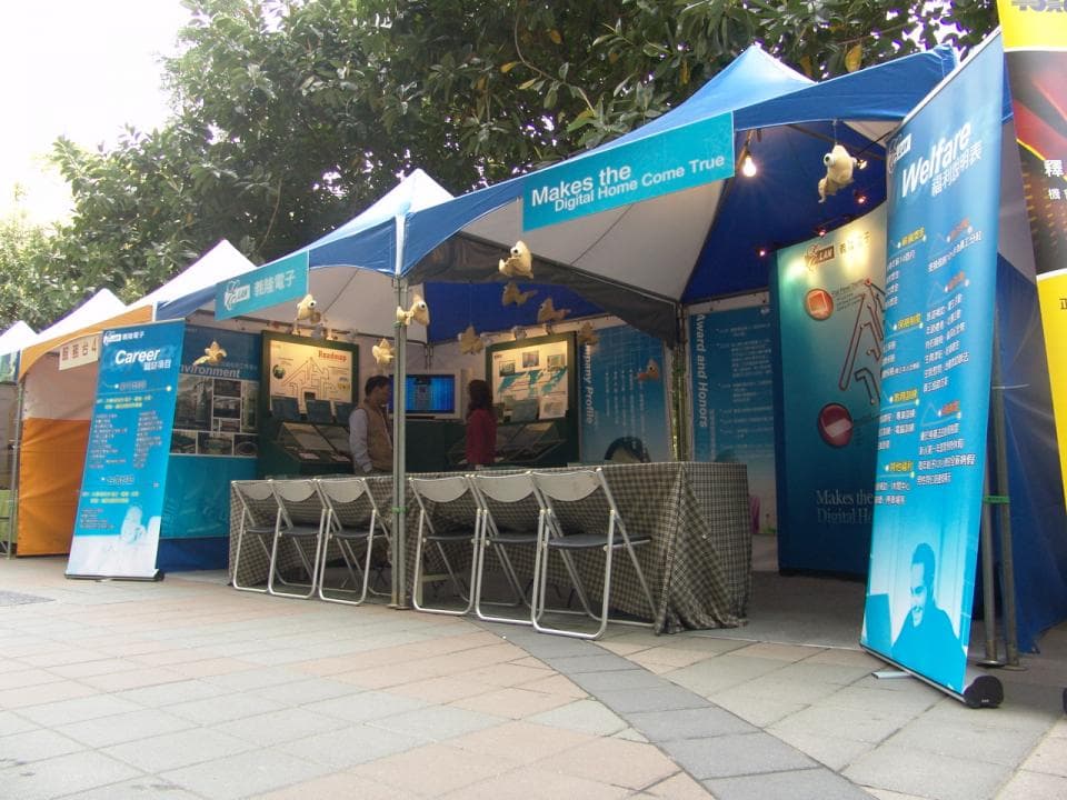 這張「戶外展覽」的照片是幾個攤位在樹下,上面寫著「Makes the Digital Home Come True」,有椅子和桌子,還有許多人。