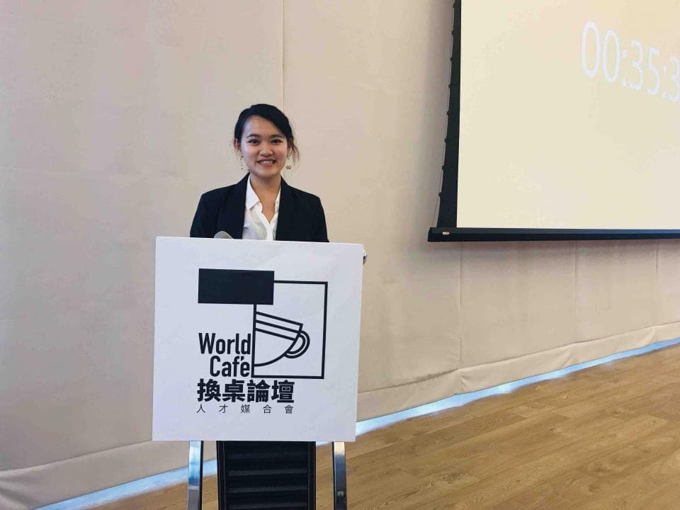 這張「世界咖啡論壇」的照片是一位年輕女性站在講臺上,她穿著西裝外套,露出笑容。背景是一面空白的投影幕,上面寫著「00:35」。
