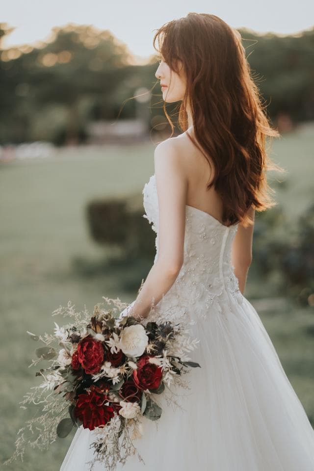 這張「婚紗照」的照片是一個新娘穿著白色婚紗,手持紅色和白色花束,站在戶外綠色草坪上,背對鏡頭,低頭看著手中的花束。