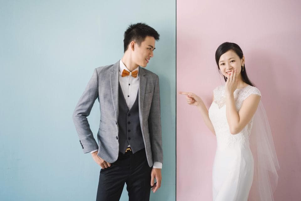 這張照片是一對新人在粉藍色和粉紅色的背景下拍照,新郎穿著灰色西裝,新婦穿著白色婚紗,他們互相看著,非常甜蜜。