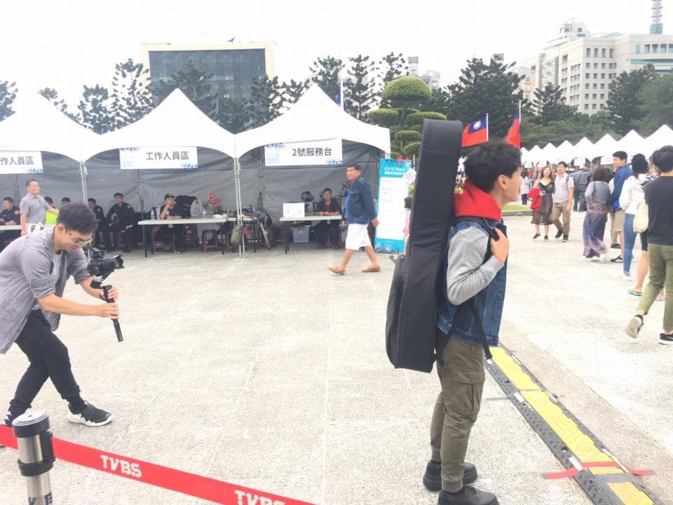 這張照片是一個音樂家,他背著一把吉他,站在一個大型的活動現場。他身穿一件牛仔外套,戴著一頂帽子,看起來很酷。