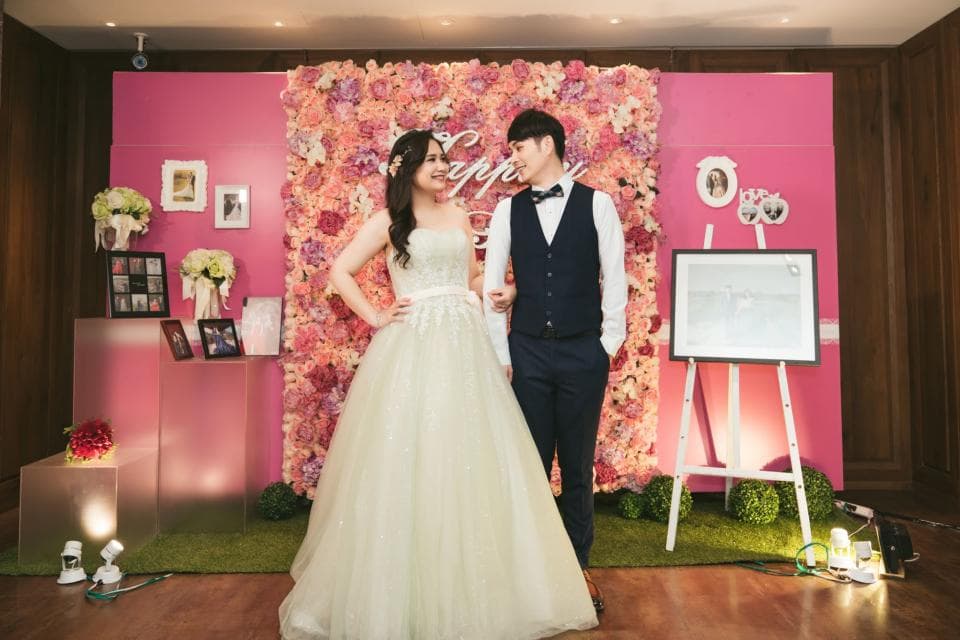 這張照片是一對新人站在花牆前,新娘穿著白色婚紗,新郎穿著黑色西裝,兩人相視而笑,幸福洋溢。