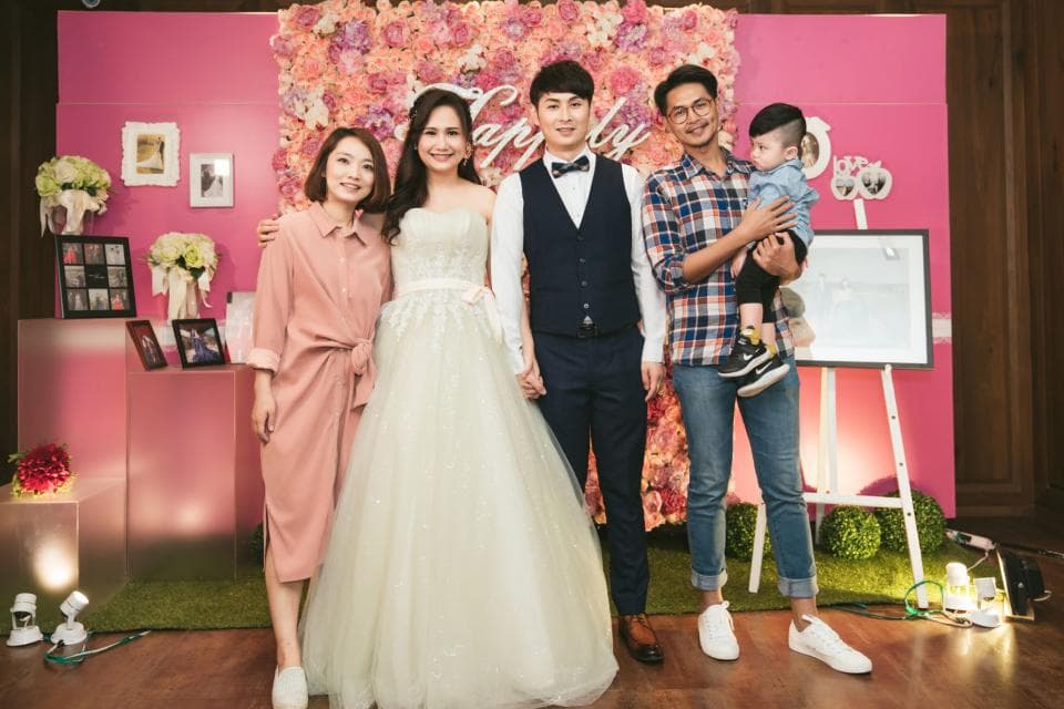 這張照片是一對新人在婚禮現場拍的合照,新郎穿著西裝,新娘穿著白色婚紗,他們身邊站著新郎的家人和朋友,其中有一個小男孩抱在其中一個朋友的懷裡。