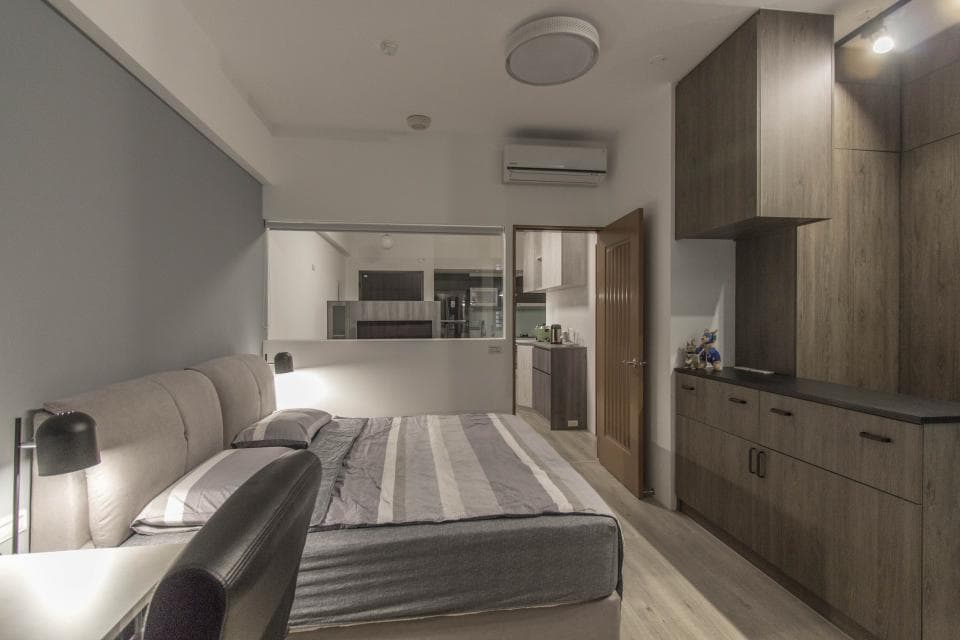 這張照片是一間現代化的臥室,最主要的部分是床鋪。床鋪上有灰白色條紋床單,舒適且典雅。床頭櫃上擺放著一盞黑色檯燈,散發出溫暖的燈光。床頭板採用柔軟的米白色布料,搭配灰色牆面,營造出舒適放鬆的氛圍。