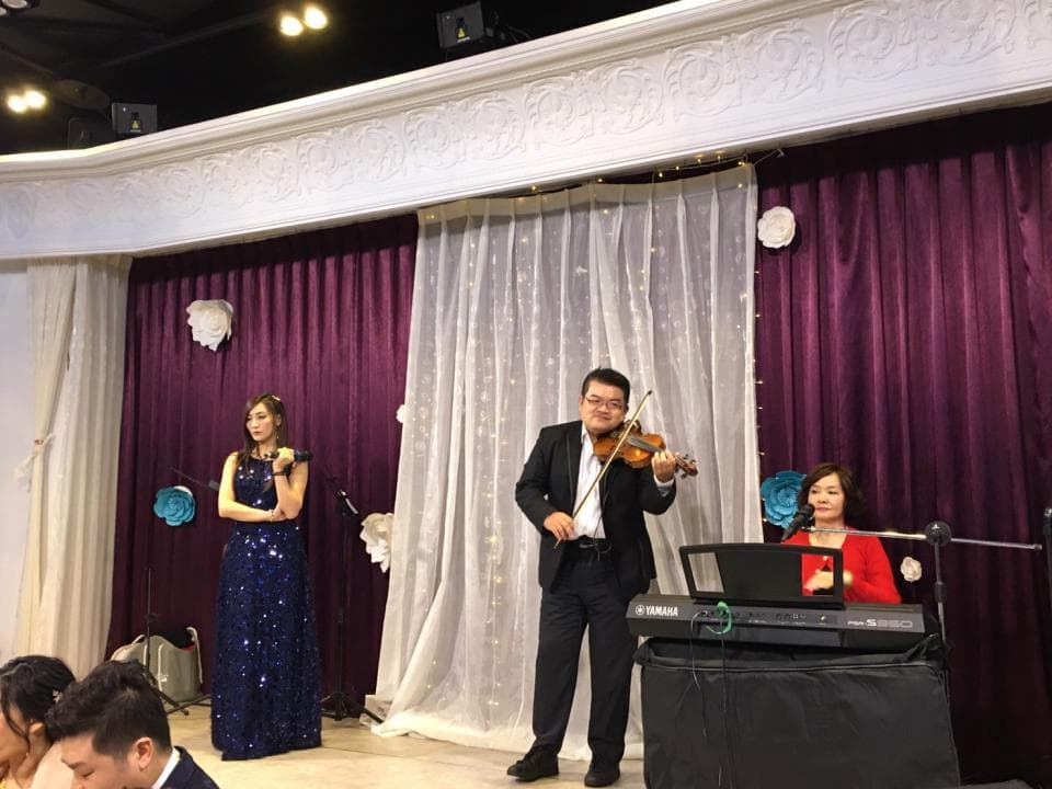 這張婚禮的照片是一個小型樂團在演奏,由一名穿著黑色西裝的男子拉小提琴,一名穿著藍色禮服的女子唱歌,以及一名穿著紅色的女子彈鍵盤。舞臺佈置簡潔,以白色蕾絲布料為主,營造出浪漫的婚禮氛圍。