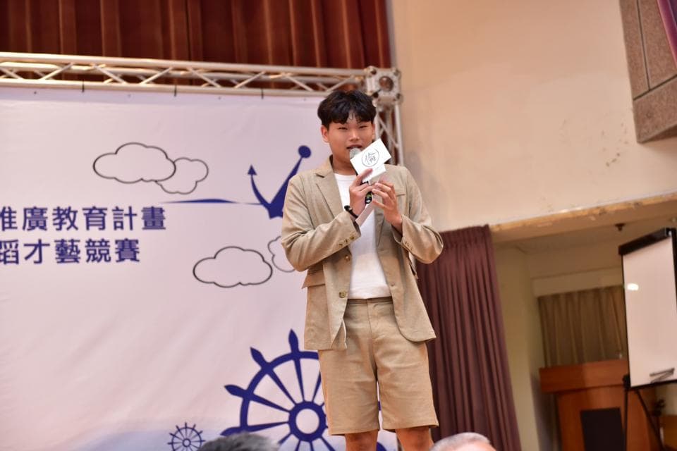 這張「一位年輕男子在臺上發表演講」的照片是,他穿著淺色的西裝外套和短褲,手持麥克風,背後是舞臺佈景,佈景上印有藍色的圖案和文字。