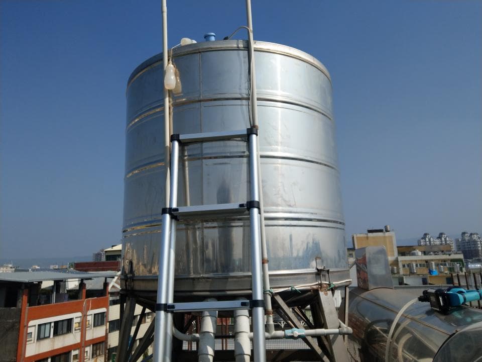 這張照片是一個大型的不銹鋼水箱,上面有一個金屬梯子,梯子似乎是為了方便清潔和維護而設計的。水箱的周圍還有其他管道和設備,可能與水系統有關。