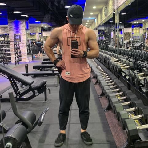 這張健身房的照片是一個穿著粉紅色背心和黑色褲子的男人,他站在一個充滿啞鈴的健身房裡,他正在用手機自拍。