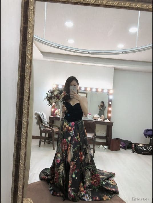 這張照片是一個穿著黑色禮服的女孩,她站在鏡子前,手拿著一束花,她的禮服上印有美麗的鮮豔花朵圖案,裙擺很長,顯得優雅動人。