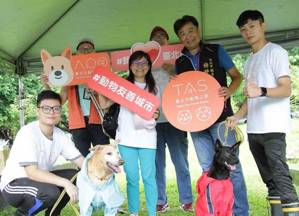 這張照片是一個在戶外舉辦的動物友善活動,照片裡有許多人以及狗狗,其中最重要的是那隻穿著紅色雨衣的狗狗,它站在眾人中間,顯得格外可愛。