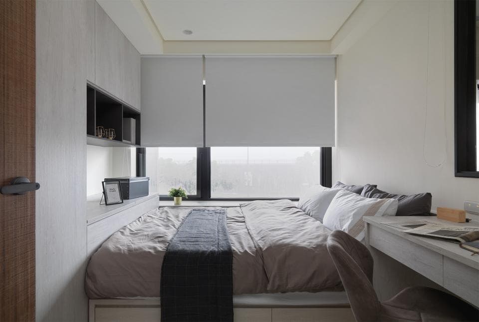 這張照片是一間臥室,最主要的部分是床鋪。床鋪上鋪著灰色床單,被子是灰色的,並且配有一條黑色的毛毯。床頭櫃上擺放著書籍和花瓶,床頭旁邊還有窗戶,可以看見外面的景色。