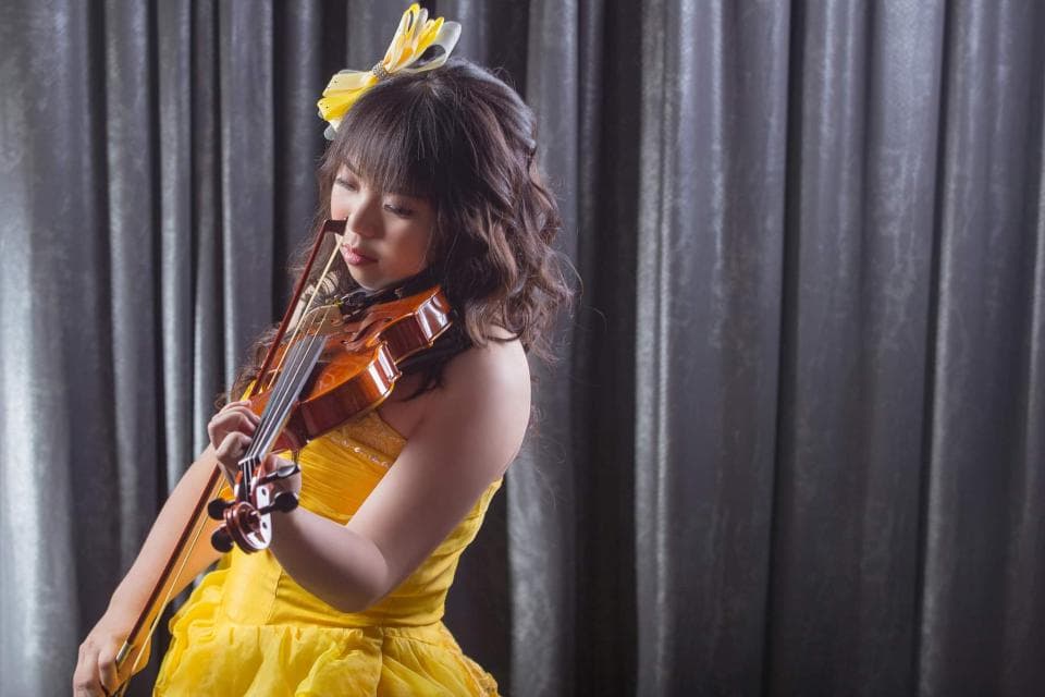 這張照片是一個年輕女子彈奏小提琴的畫面。她身穿黃色的禮服,頭上戴著黃色的蝴蝶結,專注地演奏著小提琴,看起來非常優雅。
