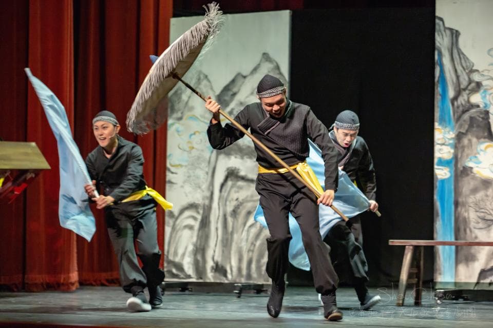 這張照片是三個穿著傳統服裝的演員在舞臺上表演。其中一位演員手持一把長柄傘,正在進行武術表演。演員們的表情和動作都充滿了戲劇性,他們正在為觀眾呈現精彩的演出。