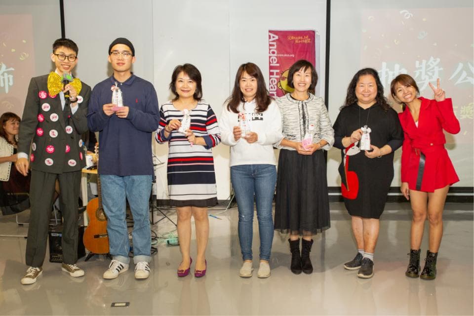 這張「一群人站在一起,拿著獎盃」的照片是,左邊第一個戴眼鏡穿著淺藍色外套的男士,右邊第一個穿著紅色的女士,還有中間的四個女士,他們都拿著獎盃,看起來很開心。