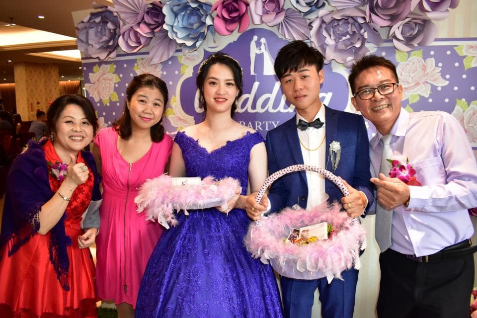 這張照片是一群人在婚禮上拍照,新郎穿著藍色的西裝,新娘穿著紫色的婚紗,他們站在一起,旁邊有伴娘和伴郎,他們都面帶笑容,看起來很開心。