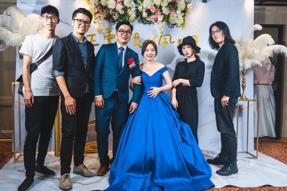 這張「婚禮」的照片是新郎新娘和朋友們在婚禮現場的合照。新娘穿著藍色的婚紗,新郎穿著藍色的西裝,朋友們則穿著休閒服裝。背景是婚禮佈置,有花朵、氣球和燈光。