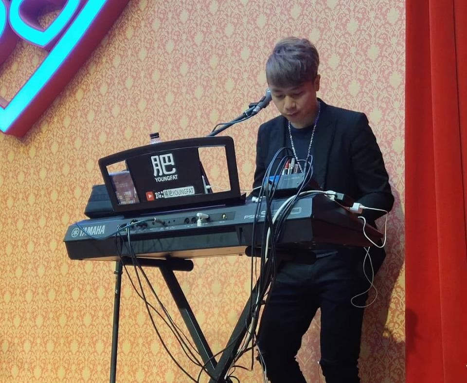 這張「一位男生在舞臺上彈奏電子琴」的照片是,他穿著黑色的西裝外套,戴著耳機,正在彈奏一個黑色的電子琴,背景是一面紅色的牆和一個金色的牆紙。