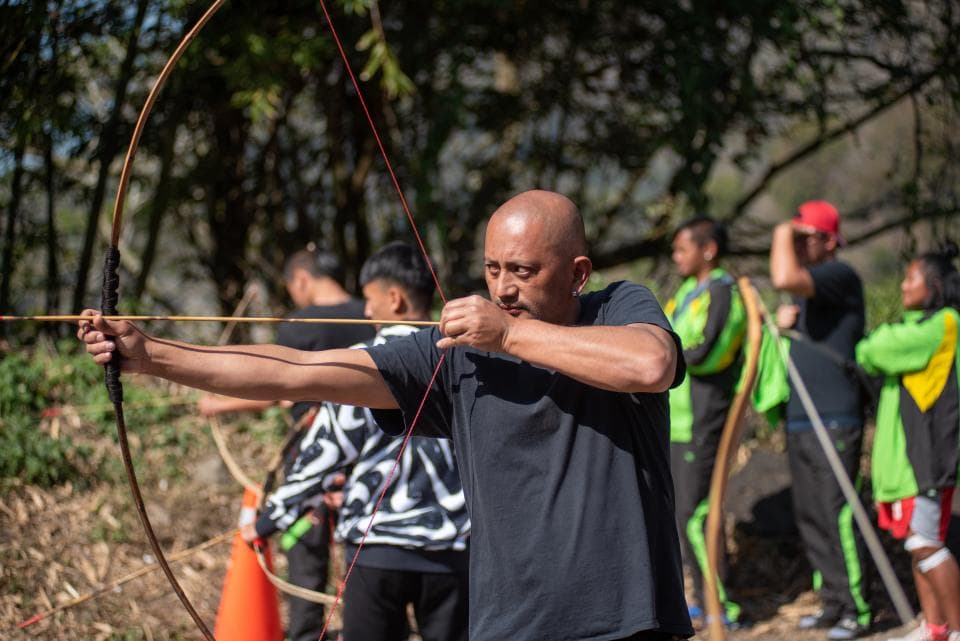 這張「射箭」的照片是一個男人正在射箭,他專注地瞄準目標,弓箭被他緊緊握住。他的背景是一片森林,還有其他人也正在練習射箭。