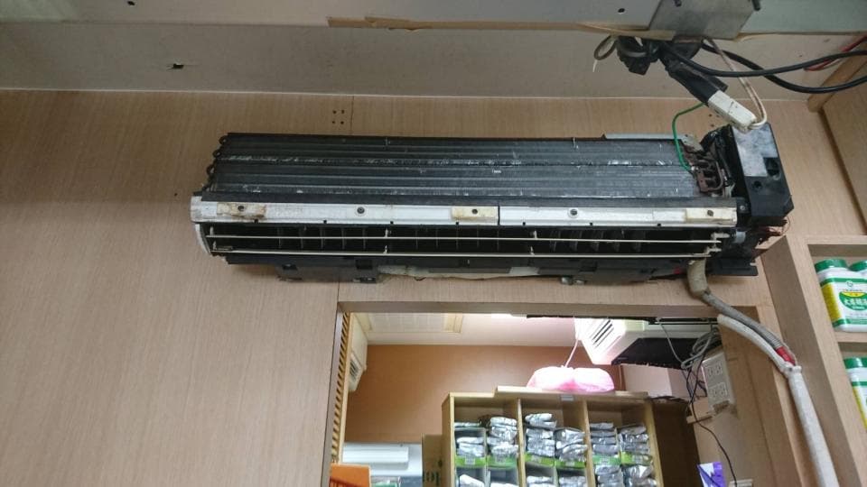 1) 照片顯示一個掛在牆上的室內機冷氣機組,冷氣機組看起來很舊,冷氣機組上方有許多電線,冷氣機組下方有木架,木架上放滿了商品