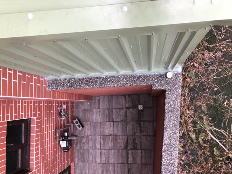 這張「房子外觀」的照片是一個綠色的屋頂,屋頂上有很多螺絲,屋頂下面是紅色的磚牆,牆上有一個窗戶,旁邊有石頭裝飾。