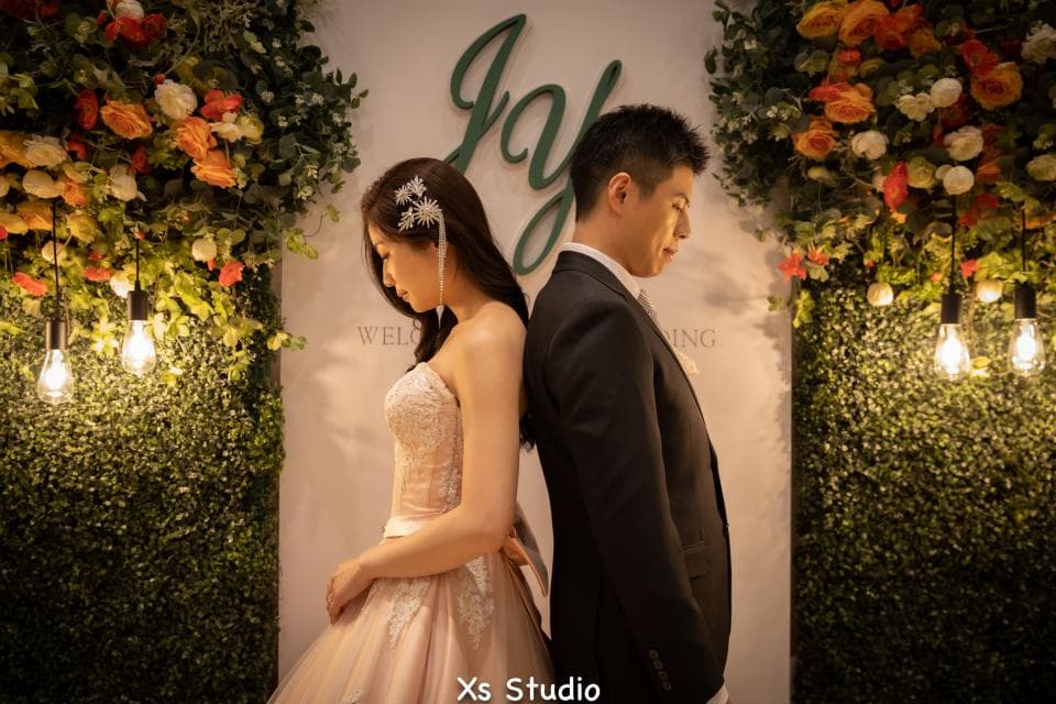 這張婚禮照片是一對新人站在綠色植物牆前,新娘穿著白色婚紗,新郎穿著黑色西裝,他們背對背站著,背景有一個寫著“Joy”的裝飾。