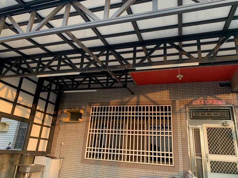 這張「遮雨棚」的照片是一個由黑色金屬製成的遮雨棚,它覆蓋在房屋的入口上方,提供遮蔽雨水和陽光的保護。遮雨棚的設計是簡單的格子狀,並帶有透明的頂棚,讓光線可以穿透。