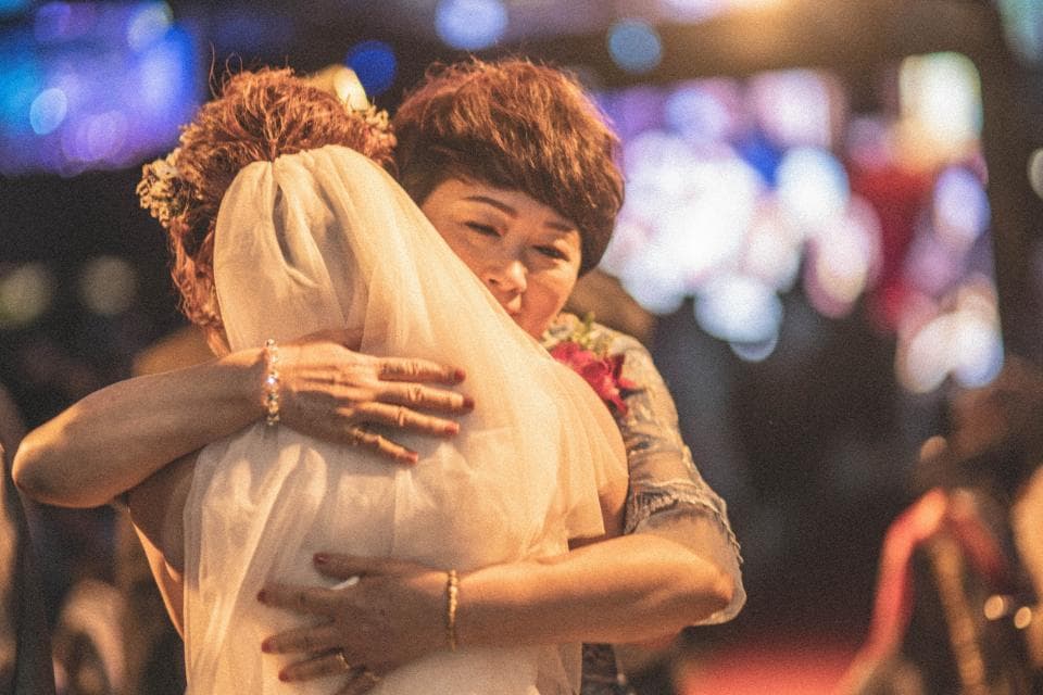 這張照片是兩位女性擁抱在一起的溫馨畫面。她們看起來很親密,新娘穿著白色婚紗,頭紗遮住了臉,而另一個女性則穿著藍色的上衣。她們互相依偎,新娘的手搭在另一個女性的肩膀上,另一個女性則緊緊地摟住新娘。她們的臉上都洋溢著幸福的笑容。