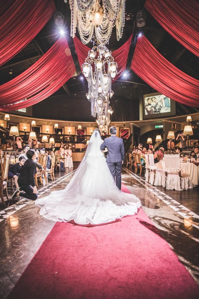 這張「婚禮」的照片是一個穿著白色婚紗的新娘,牽著穿著西裝的新郎走過紅色地毯。周圍佈置著紅色布幔,以及許多華麗的水晶燈。