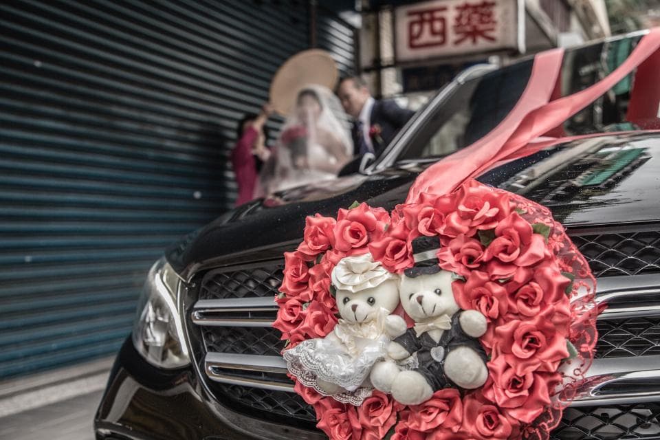 這張照片是一輛黑色轎車,車頭上裝飾著一個用紅色玫瑰花瓣做成的愛心,愛心中間還放了兩隻可愛的毛絨熊玩偶。