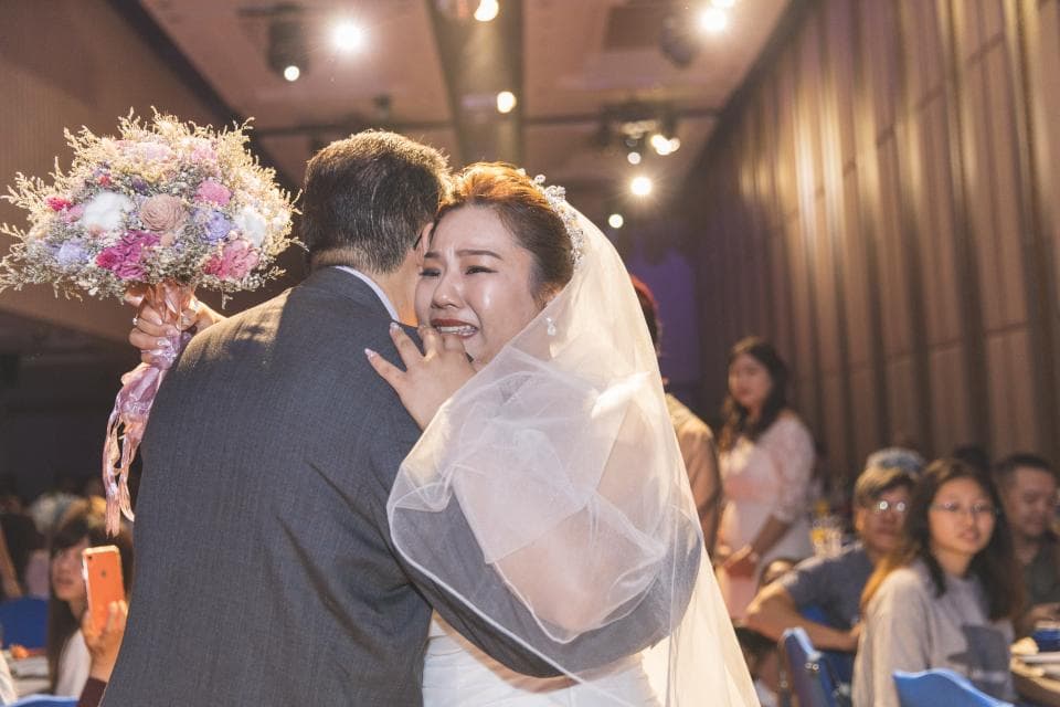 這張婚禮照片是一對新人正在擁抱,新娘穿著白色婚紗,新郎穿著西裝,新娘看起來很感動,眼泛淚光,背景是婚禮現場。