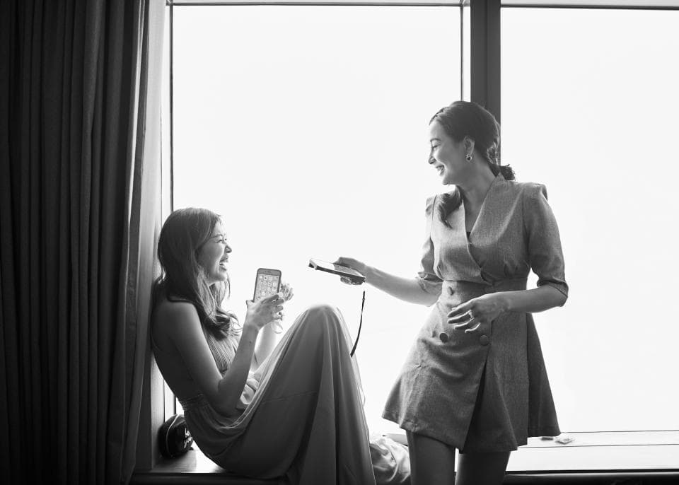 這張照片是兩個女人在窗邊聊天,其中一個女人拿著手機,另一個女人笑得很開心。她們看起來很放鬆,很享受彼此的陪伴。