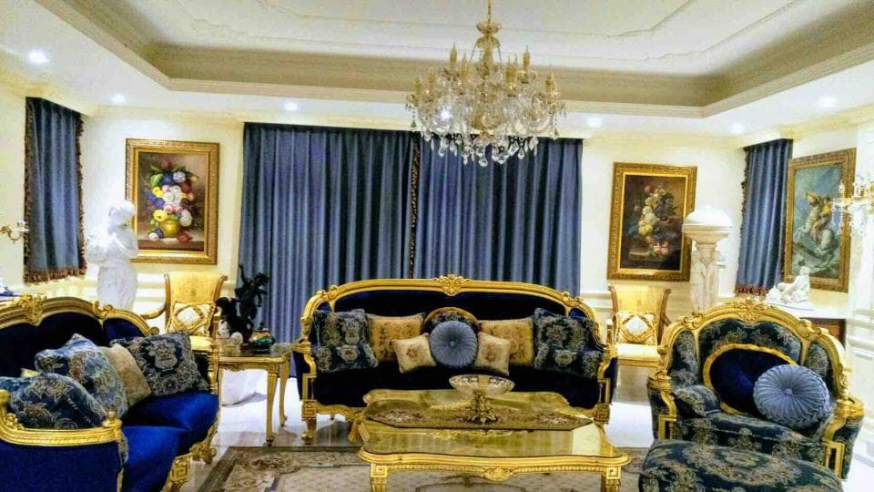 1) 照片顯示一間裝飾華麗的客廳,客廳裡有兩張金色框架的沙發,還有兩張配套的單人椅和一張咖啡桌,沙發和椅子上都有藍色的坐墊和靠墊,牆壁上有兩幅畫,窗戶上有藍色的窗簾,天花板上的水晶吊燈