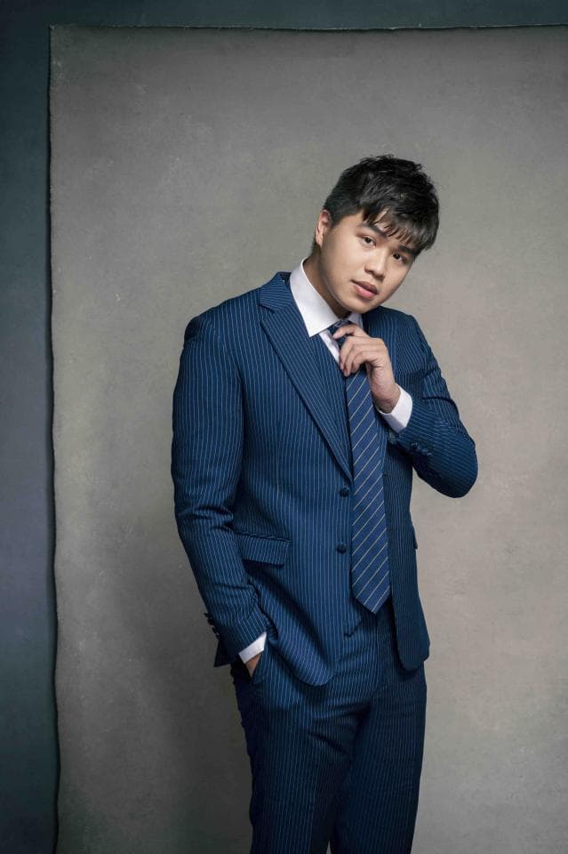 這張「穿著藍色條紋西裝的年輕男子」的照片是,他站在灰色背景前,一隻手插在口袋裡,另一隻手正在整理領帶。