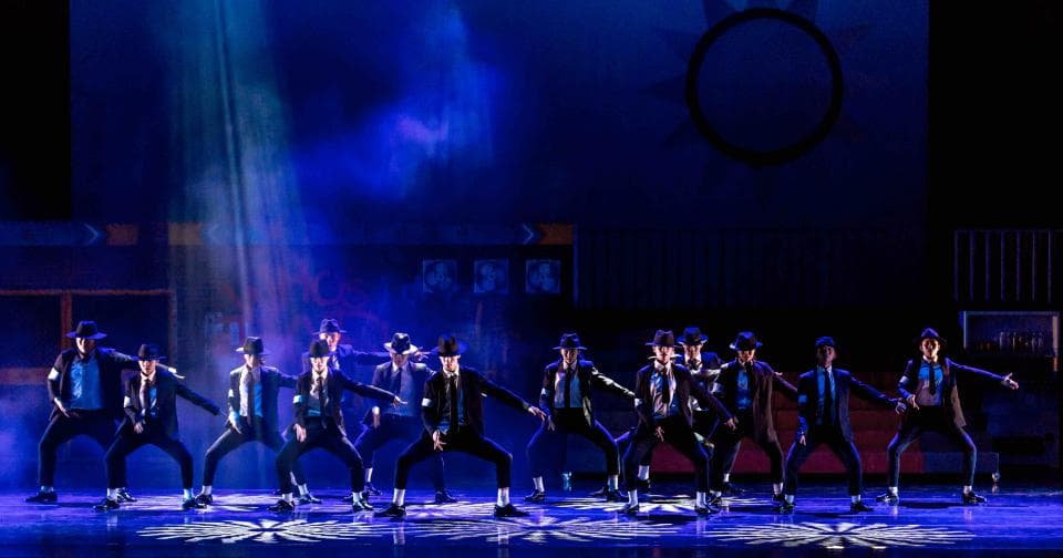 1) 照片顯示一群穿著黑色西裝的男性舞者,在黑暗的舞台上表演,舞台上裝飾著藍色燈光