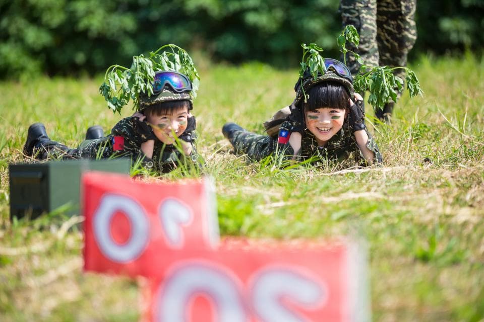 這張「兩個孩子玩遊戲」的照片是兩個孩子穿著迷彩服,頭戴綠色植物,趴在草地上,看起來很開心。
