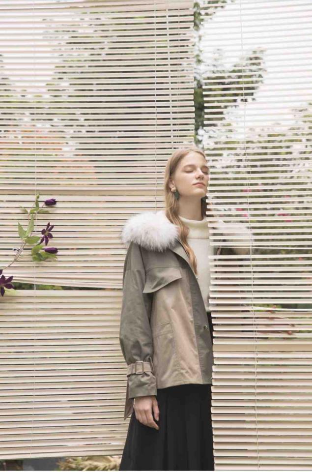 這張照片是一個年輕女子穿著一件帶有毛領的綠色外套,她站在百葉窗前,姿勢優雅,神情自然。