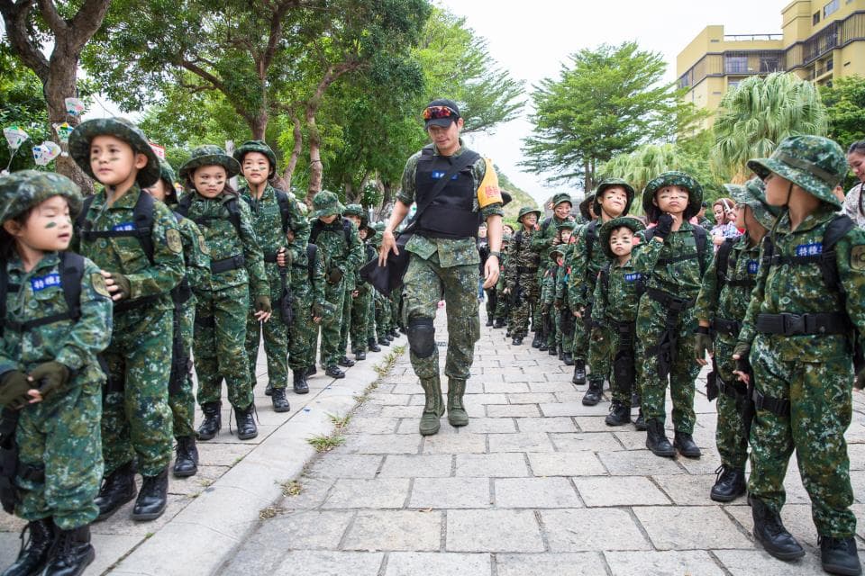 這張「一群身穿迷彩服的兒童,排成隊伍,在一位成年男子帶領下,進行戶外活動」的照片是。