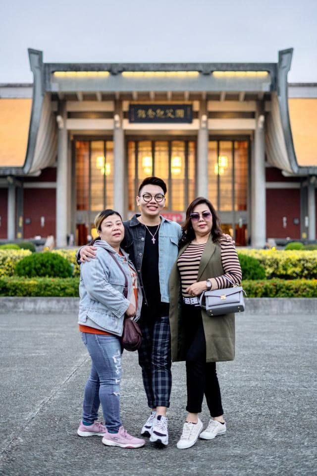 這張照片是一家人在臺灣的國父紀念館前合照。照片的重點是三個人站在一起,其中一個男生站在中間,而兩個女生站在兩邊。男生穿著牛仔外套,女生則穿著外套和連衣裙。他們看起來很開心,很享受彼此的陪伴。
